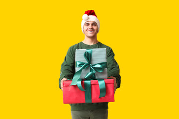 Handsome young man in Santa hat holding Christmas gifts on yellow background