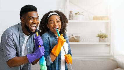 Housework with fun. Happy african couple cleaning home together, singing and fooling with mop and...