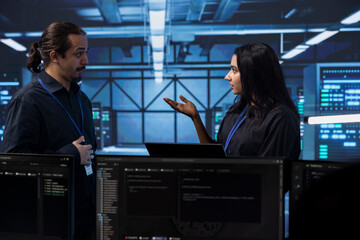 Programmers in server room discussing next steps after checking recovery plan and monitoring energy consumption. Colleagues in data center location talking about routine disk checks results