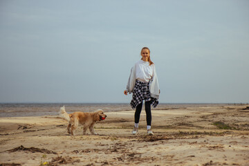 Cheerful girl on a beach walk with her dog, smiling at the camera. Warm autumn attire and relaxed seaside setting create a carefree and joyful mood.