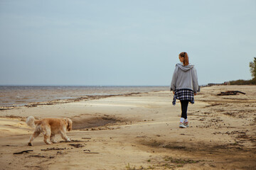 Young woman takes her dog for a peaceful beach walk on an overcast day. Casual and cozy autumn attire adds to the quiet and serene coastal vibe.