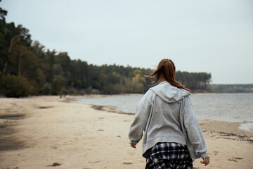 A girl strolls down the beach in a gray hoodie and a plaid shirt tied at her waist, framed by the tranquil ocean and vibrant forest, showcasing the harmony of style and natural beauty.