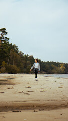 Walking along the sandy beach, a girl dressed in a cozy gray hoodie and a tied plaid shirt enjoys the stunning view of the sea and lush forest, capturing a perfect moment in nature. 