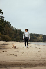A girl in a gray hoodie and plaid shirt tied around her waist strolls along an empty beach with her light-colored Cocker Spaniel, enjoying the autumn breeze and the sound of the waves. Vertical photo