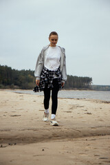A girl in a gray sweatshirt and a black-and-white plaid shirt tied around her waist strolls along the beach, with the ocean waves and lush forest creating a picturesque backdrop. Vertical photo
