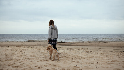 Woman and dog taking a relaxed walk along the beach. Dressed for autumn, with a plaid shirt tied at the waist. Coastal outdoor activity emphasizing the bond between human and pet.