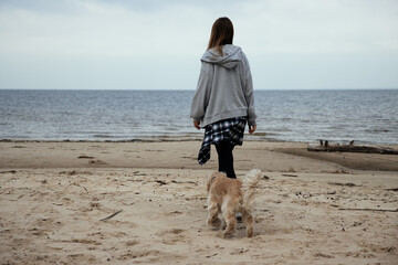 Woman enjoying a beach walk with her dog, dressed in cozy autumn clothes. Plaid shirt around her waist adds a relaxed, casual vibe. Ocean shore in the background.
