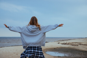 Young woman walking on a sandy beach with her arms outstretched, embracing the feeling of freedom and nature. Serene autumn day by the sea, symbolizing peace and liberation.