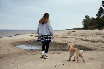 Woman walking her dog along the shore, wrapped in warm autumn clothing. The serene beach setting creates a peaceful outdoor mood, perfect for themes of nature and pet care.