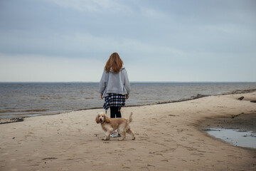Girl walking her dog along the beach on a cloudy day. Casual autumn outfit with a plaid shirt tied around her waist. Outdoor recreation with a pet by the sea.