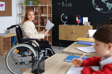 Mature teacher in wheelchair with clipboard conducting Geography lesson to little pupils at school