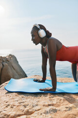 Woman practicing yoga on a blue mat by the sea at sunrise She wears headphones and focuses on her pose, surrounded by natural rock formations