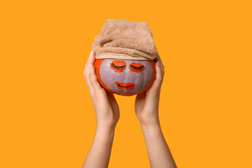 Woman holding funny pumpkin with cosmetic mask and towel on yellow background, closeup