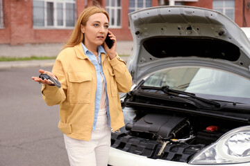 Mature woman with key talking by mobile phone near broken car on city street