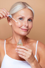 Skincare beauty portrait of a mature senior woman with a grey silver hair and glowing skin applying a facial care serum or retinol with a pipette dropper against beige background.