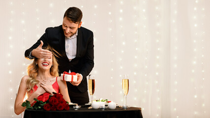 Romantic man surprising his girlfriend with gift on date in restaurant, covering her eyes and...
