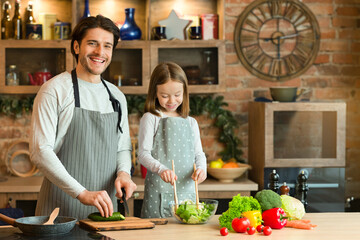 Happy father and daughter cooking healthy breakfast, lunch, or dinner together in kitchen, enjoying organic food, preparing vegetable salad in bowl, wearing aprons, having fun at home, copy space