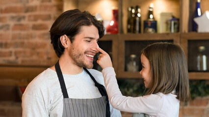 Happy little girl having fun with her dad while cooking together in kitchen, playfully rubs his nose with flour and laughing