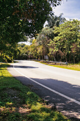 The country road stretches gently through a vibrant landscape in Melaka, Malaysia, lined with trees under a clear blue sky, creating a peaceful atmosphere perfect for a leisurely drive.
