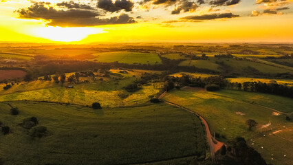 Fototapeta premium Imagem aérea de um campo rural ao pôr do sol, com colinas e áreas verdes iluminadas por tons dourados. Ideal para temas de agricultura, natureza e paisagens serenas.