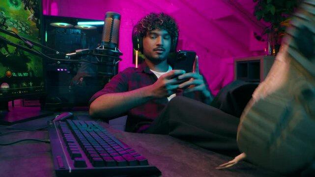Indian Muslim man gamer streamer influencer in headphones put feet on table using mobile phone smartphone cyber sport player computer online video game cyberspace addict radio guy male typing keyboard