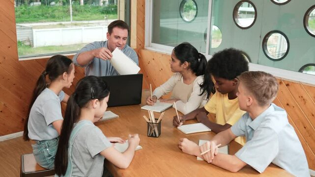 Caucasian teacher and smart boy discuss about project at classroom. Professional instructor talking about classwork while giving a paper to high school child with diverse student listen. Edification