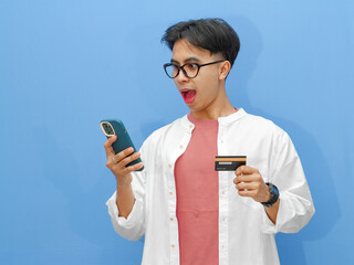 Portrait of an excited Asian man holding a credit card, using a mobile phone for online payment, celebrating success on a blue background.