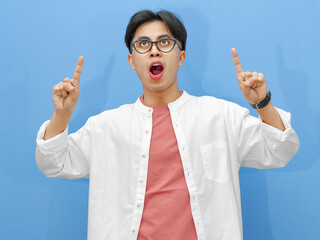 Portrait of an excited Asian man in casual clothes pointing up, showing joy and energy on a blue...