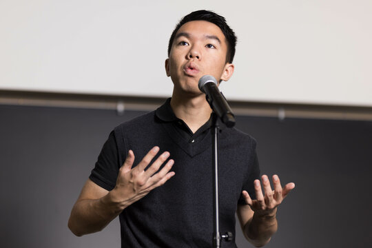 A young man confidently speaks to an audience in front of a microphone