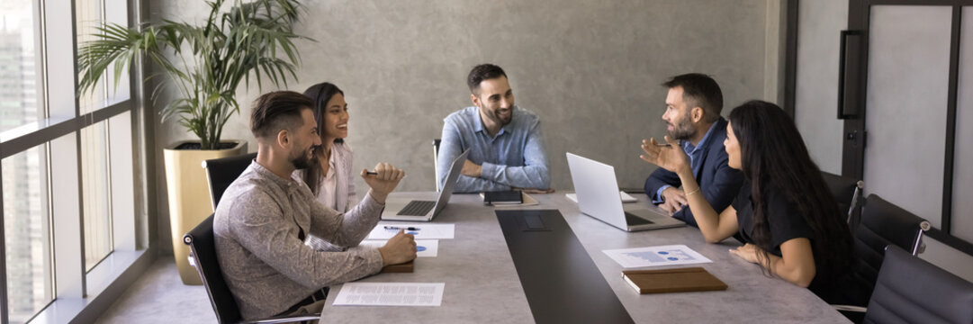 Diverse team of project managers enjoying teamwork, partnership, sitting at large conference table, talking, discussing creative work strategy, planning tasks, laughing. Banner shot