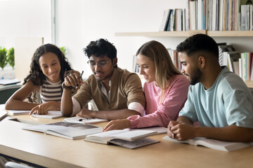 Smart diverse college students preparing scientific learning report in library together. Multiethnic group of classmates reading textbook, book at large table aloud, smiling, laughing