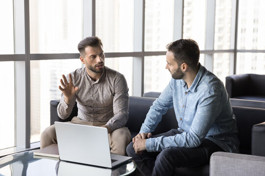 Serious busy professional man talking to business colleague, moving hands for explaining. Two entrepreneurs men meeting in office co-working hall, sitting on sofa, discussing teamwork, collaboration