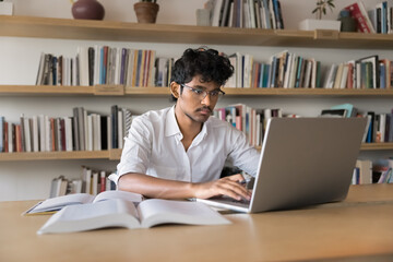 Serious young Indian student man in glasses typing on laptop in public college library, working at table with open books, textbooks, preparing report, learning lecture for class, working on research