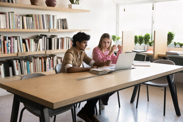 Multiethnic couple of students discussing study project in college library, talking at laptop in educational space for group work, sitting at computer on table, watching learning video tutorials