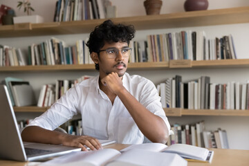 Thoughtful clever Indian student man in glasses writing scientific article, research paper in...