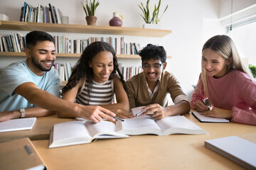 Positive focused university classmates reading learning books together in library, working on group report, enjoying teamwork, preparing for exam, talking, smiling, laughing, pointing at textbook page