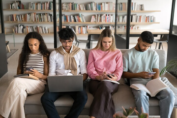 Diverse group of silent focused serious students sitting close together on couch in library, using digital gadgets, keeping silence, browsing online learning information, ignoring each other