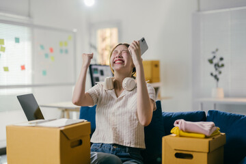 Young asian woman is raising her fist and smiling broadly while sitting between cardboard boxes, celebrating a success on her smartphone