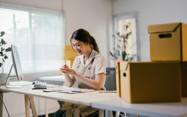 Young asian entrepreneur writing down notes while managing her business from home office, surrounded by cardboard boxes