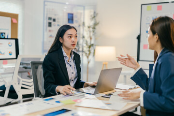 Two young businesswomen are working together on a new project, discussing ideas and analyzing data on a laptop in a modern office