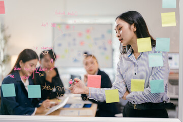 Asian businesswoman is using a pink marker to write on a glass wall during a meeting with her colleagues. The glass is covered with sticky notes