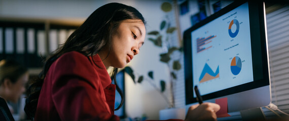 Focused businesswoman works late in an office, reviewing data on her computer screen. The scene...