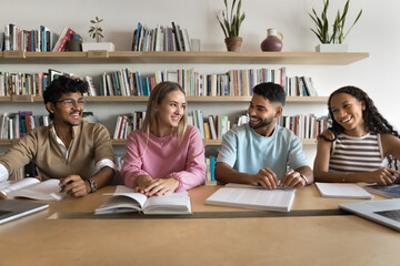 Diverse team of happy college students meeting in library, talking at large table over open books, having fun, smiling, laughing, discussing university education, studentship, community