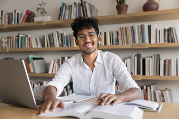 Cheerful young Indian college student man in eyeglasses sitting at table with heap of books in...
