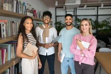 Multiethnic team of happy young college students posing in university library, standing at bookshelves, looking at camera, smiling, promoting diversity in higher education. Group portrait