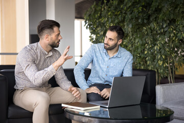 Two business partners discussing work project at laptop, sitting on couch in co-working, talking,...