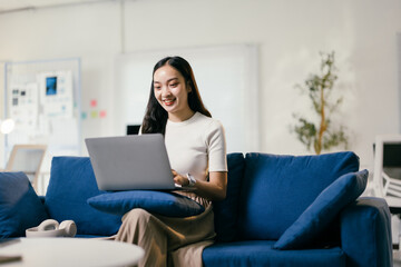 Smiling young woman is working remotely from her home office, comfortably sitting on a blue sofa while typing on her laptop