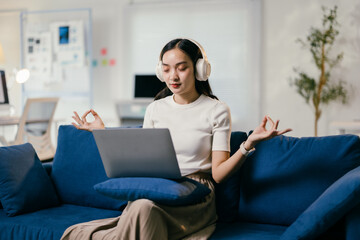 Young woman is sitting cross-legged on a sofa, meditating with her eyes closed. She is wearing headphones and listening to music on her laptop