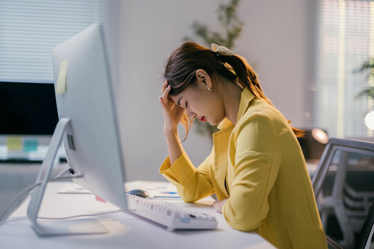 Young professional woman is sitting at her desk with her head in her hand, feeling unwell and experiencing a headache