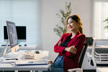Young businesswoman is sitting at her desk in a modern office, smiling with her arms crossed. She...